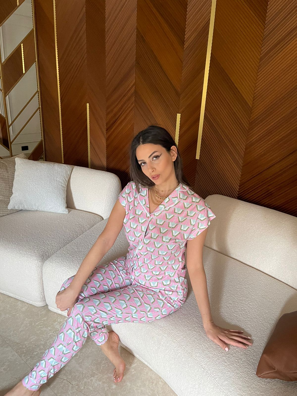 Model wearing a Popjammies pink geometric pyjama set, featuring a short-sleeve button-down top and matching pants, sitting on a beige couch with a wooden patterned background.