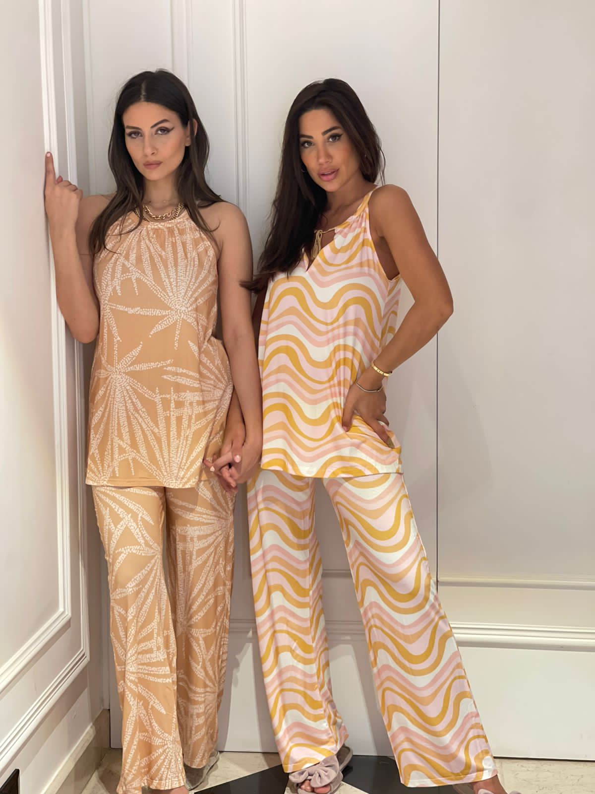 Two models standing side by side, one wearing a beige pyjama set with a white leaf pattern and the other wearing a pastel pink and yellow wave pattern pyjama set from Popjammies, posing against a white wall and holding hands, with one model slightly leaning on the wall.