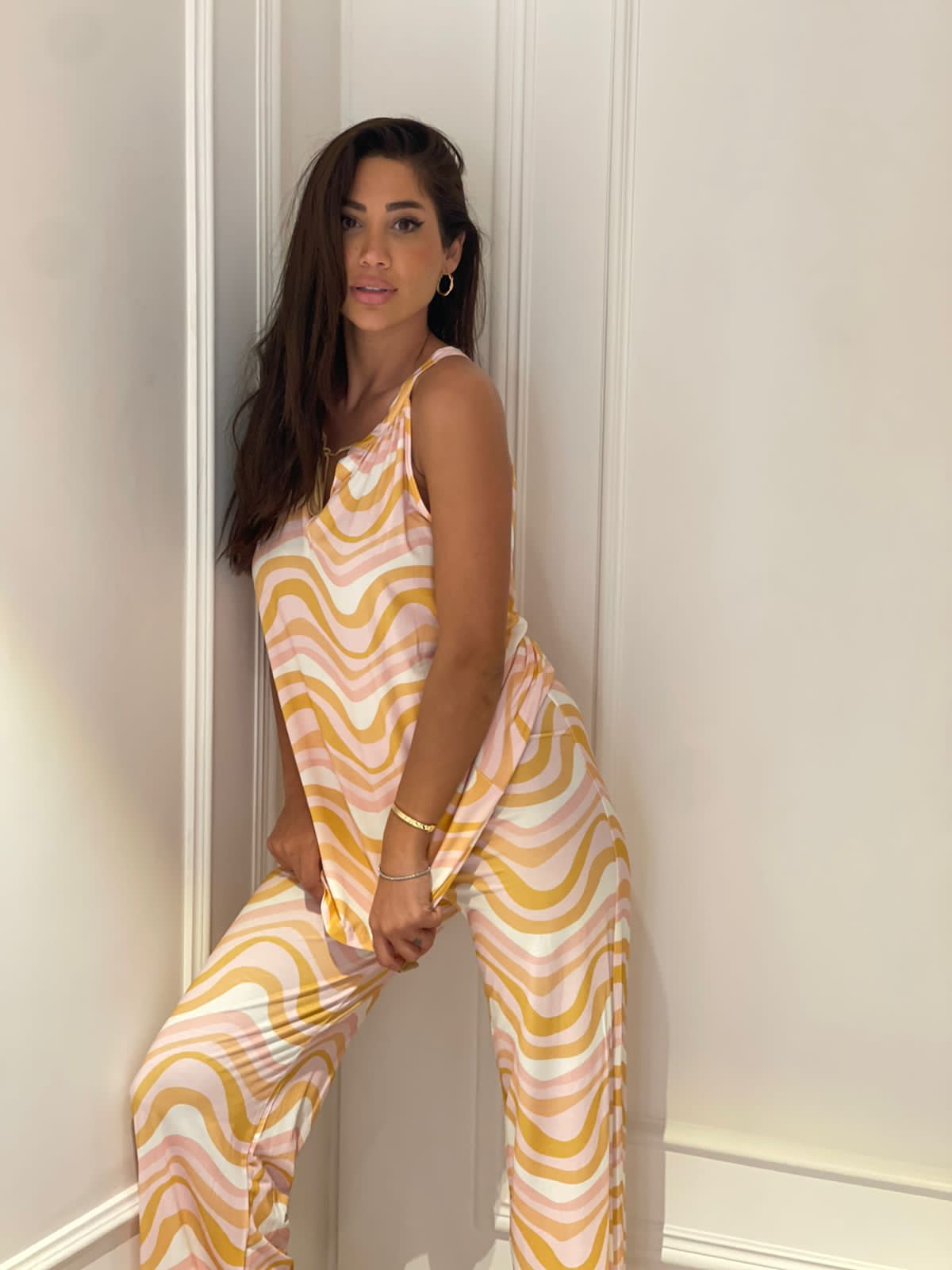 A model wearing a pastel pink and yellow wave pattern pyjama set from Popjammies, leaning against a white wall with her left hand holding the top of her pyjama pants, looking directly at the camera.
