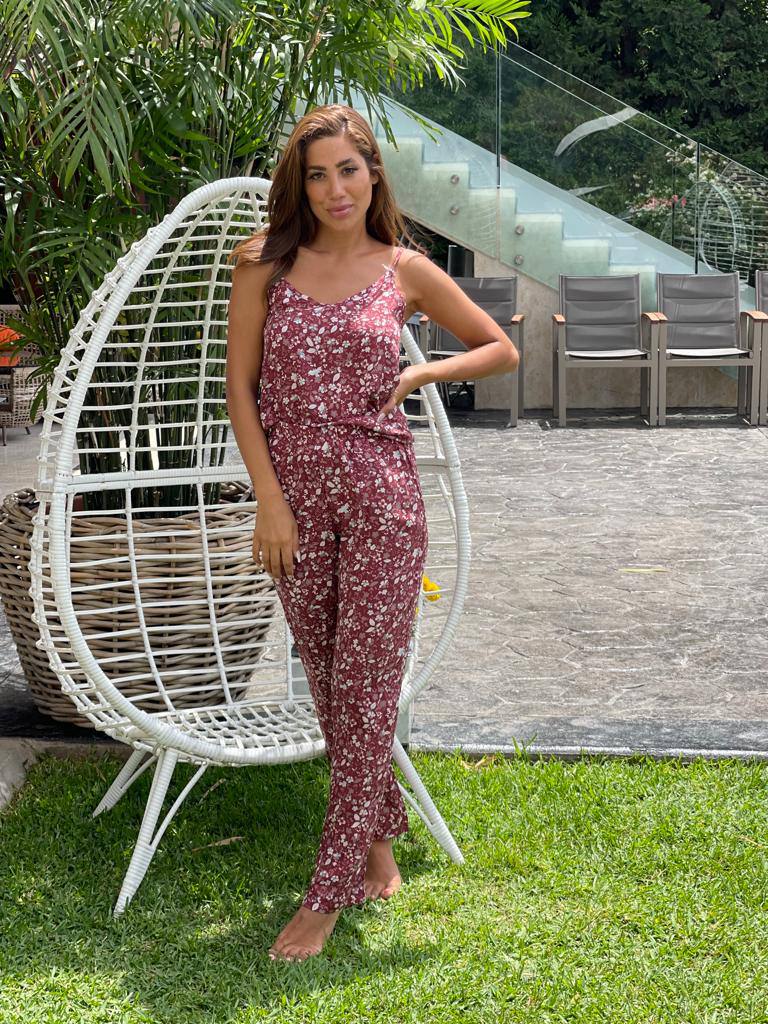 Woman standing outdoors on a grassy area, wearing a deep red floral printed pyjama set from Popjammies, with white wicker chair in the background.
