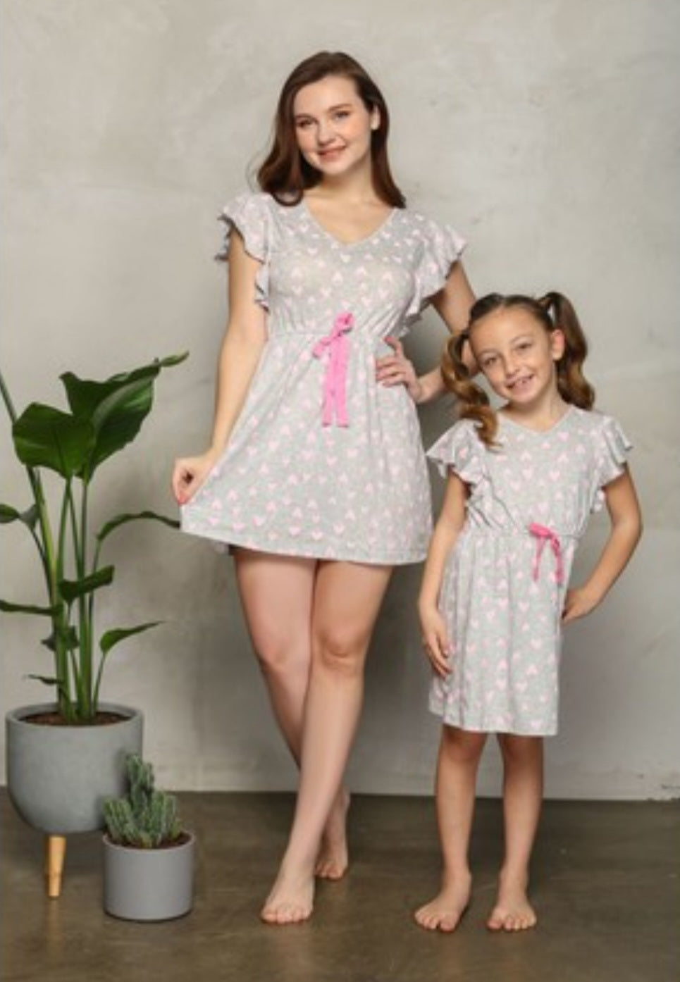 Child and mother wearing matching grey dresses with heart-shaped dots and pink ribbons, standing together in front of a white background.
