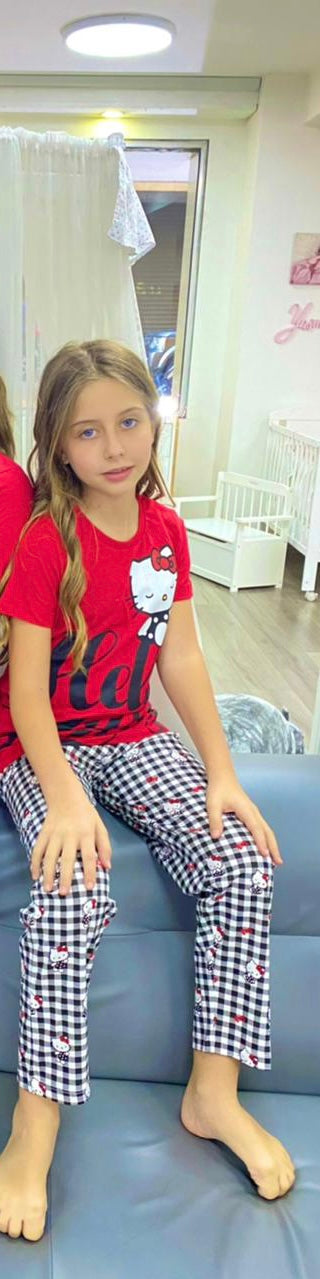A young girl sitting on a blue sofa, wearing a red Hello Kitty top and black and white checkered pyjama pants, in a brightly lit room with a white crib in the background.